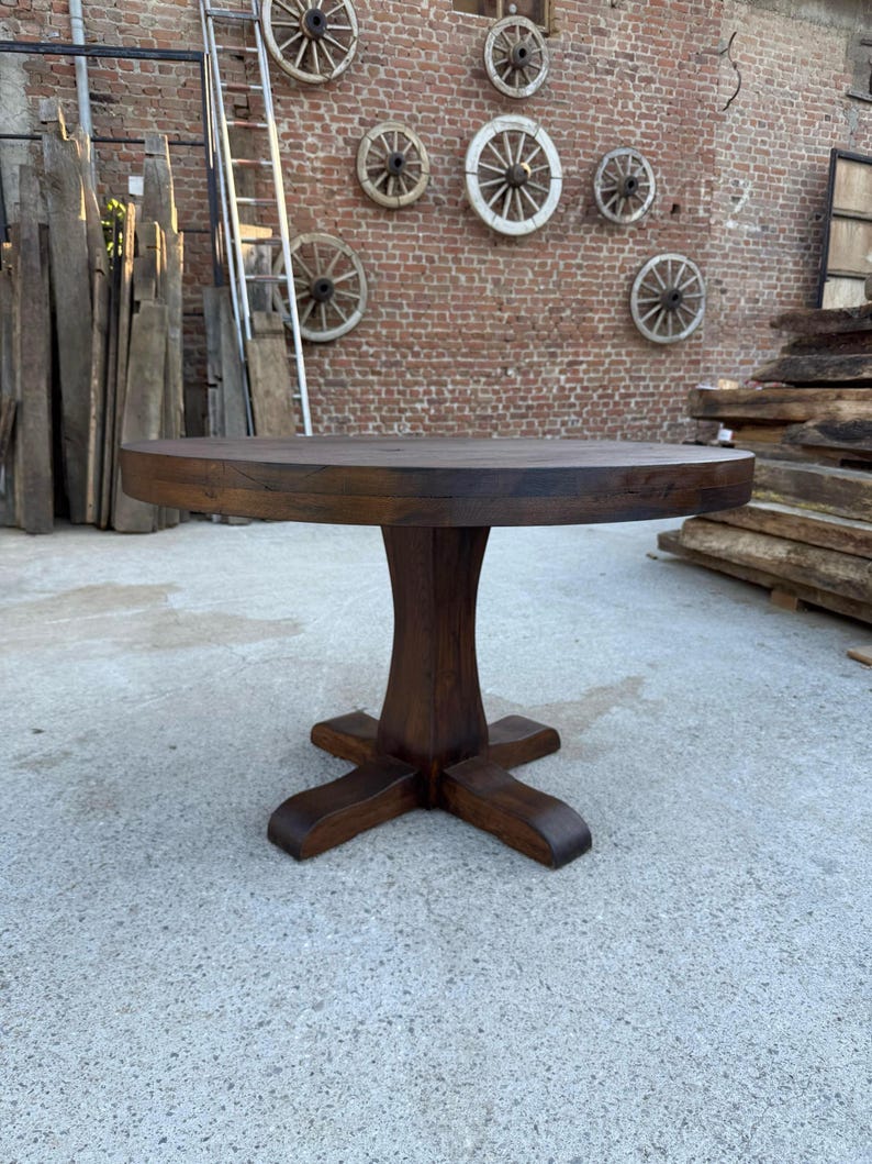 May include: A round dark brown wooden table with a thick tabletop and a central pedestal base featuring four curved feet. The table has a smooth, polished finish and a sturdy construction, standing on a flat, light-colored concrete surface.