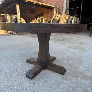May include: A round dining table crafted from dark brown wood, featuring a thick circular tabletop supported by a central pedestal base that branches out into four curved feet at the bottom, showcasing a visible wood grain texture throughout.