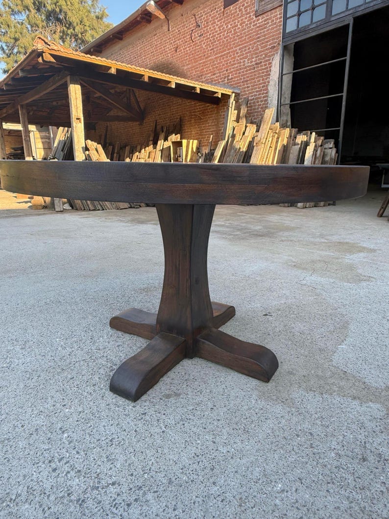 May include: A round dining table crafted from dark brown wood, featuring a thick circular tabletop supported by a central pedestal base that branches out into four curved feet at the bottom, showcasing a visible wood grain texture throughout.