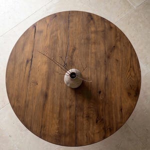 May include: A round wooden table with a visible grain pattern and a natural brown finish. Centered on the table is a small, round, light-colored ceramic vase containing a few thin, bare, dark brown twigs that extend outward from the opening.