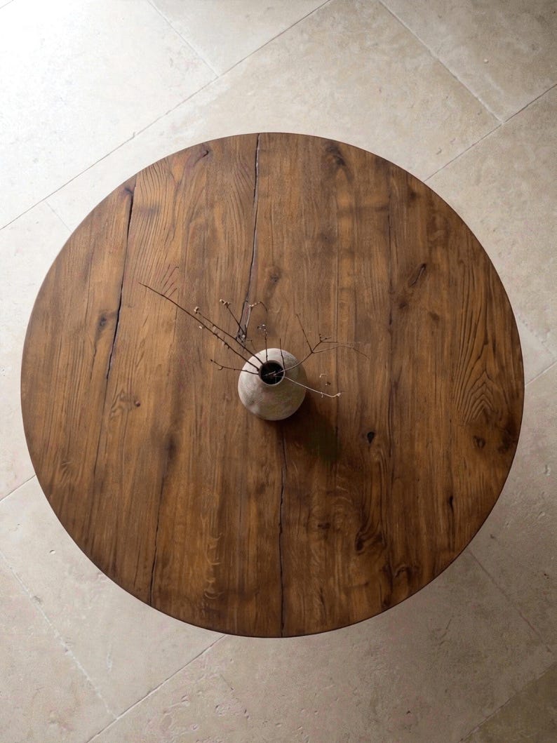 May include: A round wooden table with a visible grain pattern and a natural brown finish. Centered on the table is a small, round, light-colored ceramic vase containing a few thin, bare, dark brown twigs that extend outward from the opening.