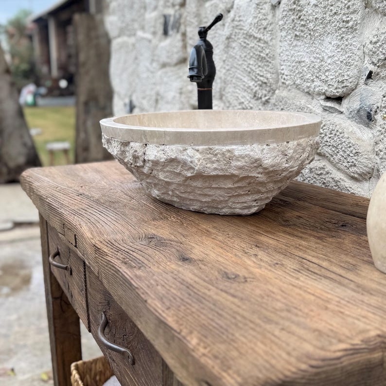 May include: A rustic wooden vanity with two drawers and a stone vessel sink. The sink is oval shaped and has a rough, textured surface. A black faucet is mounted on the wall above the sink.
