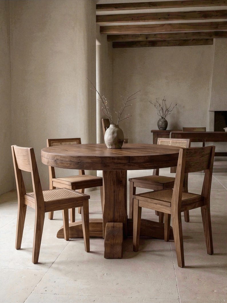 May include: A round wooden dining table with a thick pedestal base and four matching wooden chairs featuring woven cane seats. A light-colored, textured ceramic vase containing thin, bare branches sits in the center of the table surface.