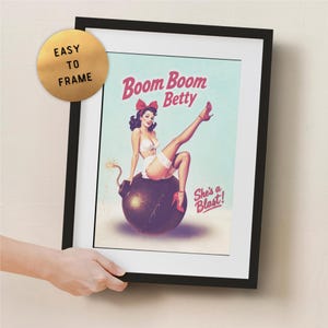 May include: Framed vintage-style poster featuring a pin-up girl sitting on a bomb. The poster has the text "Boom Boom Betty" and "She's a Blast!" in red. The frame is black, and the poster is easy to frame.