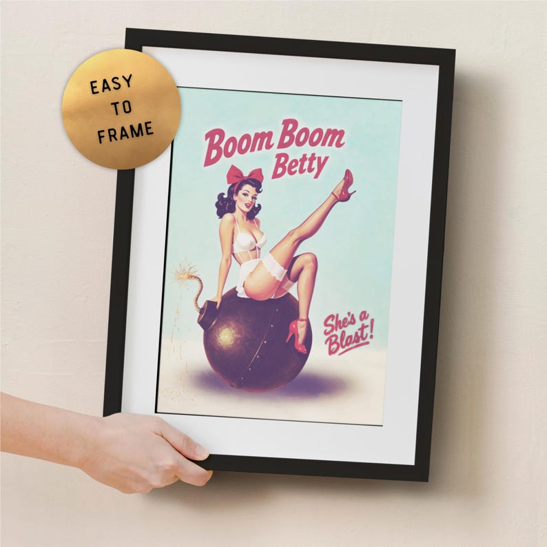 May include: Framed vintage-style poster featuring a pin-up girl sitting on a bomb. The poster has the text "Boom Boom Betty" and "She's a Blast!" in red. The frame is black, and the poster is easy to frame.
