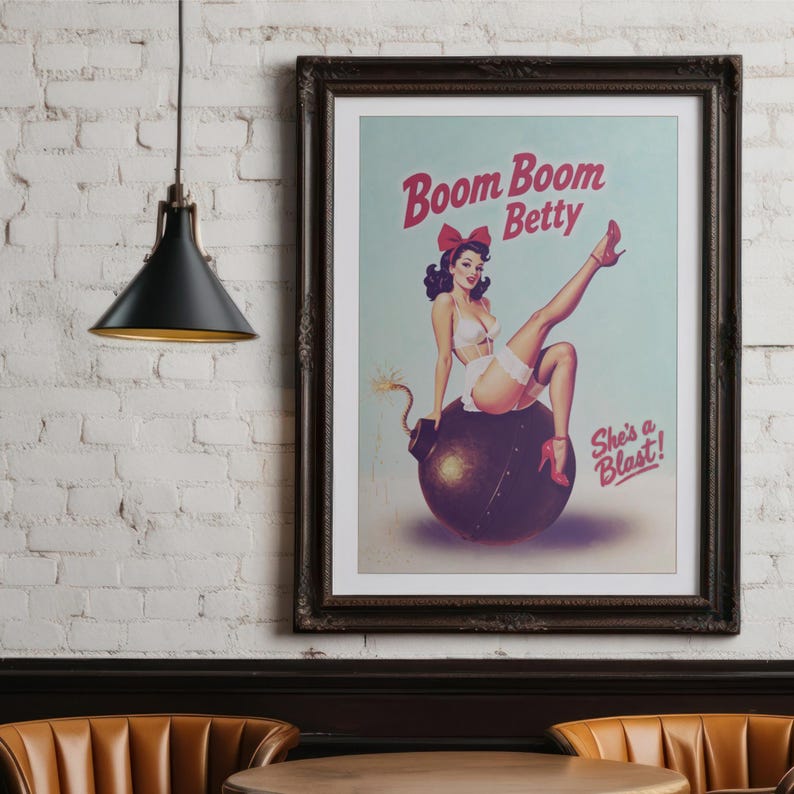 May include: Framed vintage-style poster featuring a pin-up girl sitting on a bomb. The poster has the text "Boom Boom Betty" and "She's a Blast!" in red. The image is in a dark wooden frame, hanging on a white brick wall.