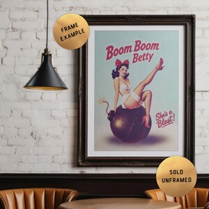 May include: Framed vintage-style print featuring a pin-up girl sitting on a bomb. The print has the text "Boom Boom Betty" and "She's a Blast!". The print is in a dark wooden frame and is displayed on a white brick wall.