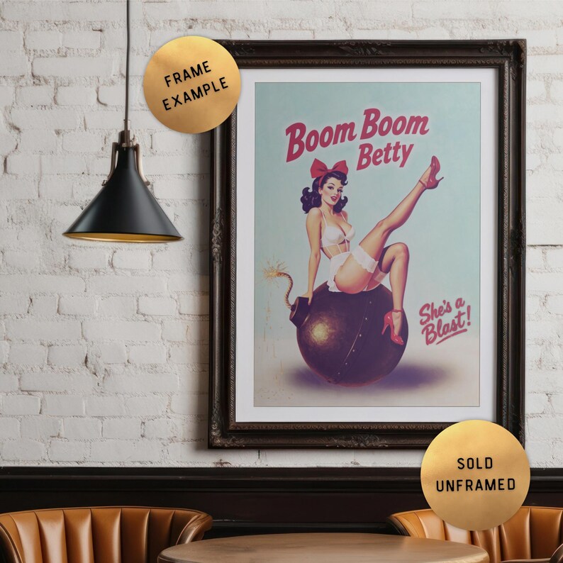 May include: Framed vintage-style print featuring a pin-up girl sitting on a bomb. The print has the text "Boom Boom Betty" and "She's a Blast!". The print is in a dark wooden frame and is displayed on a white brick wall.