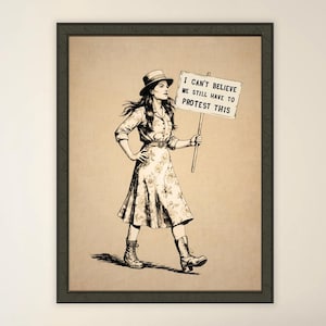 May include: Framed monochrome print depicting a woman marching and holding a sign. The sign reads, "I CAN'T BELIEVE WE STILL HAVE TO PROTEST THIS." The woman wears a hat, dress, and boots.