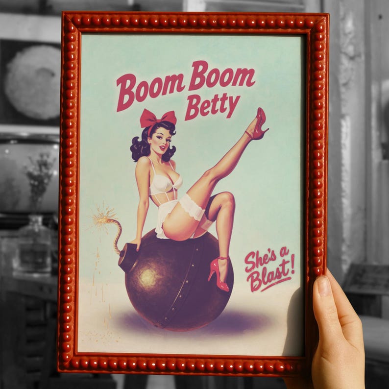 May include: Framed vintage-style print with a pin-up girl illustration. The artwork features a woman in lingerie sitting on a bomb, with the text "Boom Boom Betty" and "She's a Blast!" in red. The frame is red.