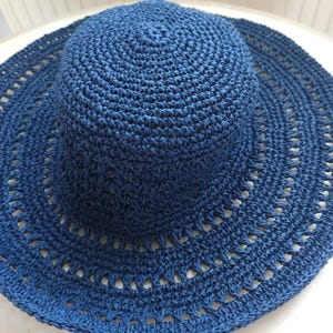 Könnte beinhalten: Ein marineblauer Sonnenhut mit breiter Krempe. Der Hut besteht aus gewebtem Material mit einer kreisförmigen Krone und einem Rand mit dekorativen Ausschnitten. Der Hut soll vor der Sonne schützen.