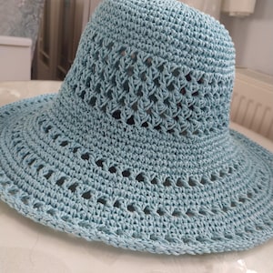 Könnte beinhalten: Hellblauer Häkel-Sonnenhut mit breiter Krempe und dekorativem Lochmuster. Der Hut hat eine runde Krone und ist aus weichem, strukturiertem Garn gefertigt. Der Rand hat einen gewellten Rand.