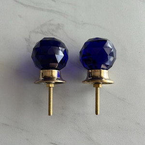 Op de afbeelding: Twee facetgeslepen, diepblauwe glazen kastknoppen met messing voetjes en schroefdraad. De knoppen hebben een geometrisch ontwerp. Deze decoratieve knoppen zijn ideaal om een vleugje elegantie aan meubels toe te voegen.