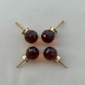 以下が含まれることがあります： 真鍮金具付きの、4つのファセットアンバーガラスキャビネットノブ。ノブはニュートラルな背景に対して正方形のパターンに配置されています。各ノブは装飾的な幾何学模様のデザインです。