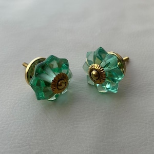 Puede incluir: Dos tiradores decorativos para armarios. Cada tirador presenta un diseño de vidrio verde esmeralda facetado con una base de metal dorado. Los tiradores tienen forma de estrella y están diseñados para una fácil instalación.