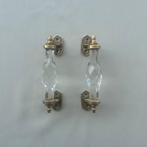 Può includere: Una coppia di maniglie per porta verticali con sezioni centrali in vetro trasparente sfaccettato con motivo a taglio di diamante. Ogni maniglia è fissata da due piastre di montaggio in metallo color ottone antico in alto e in basso.