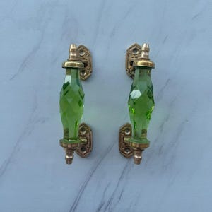 May include: Two matching cabinet pulls featuring faceted, translucent lime green glass handles. Each pull is secured by polished brass hardware with ornate, rectangular mounting plates at the top and bottom, showcasing a vertical, geometric design.