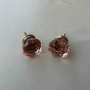 May include: Two faceted, light pink glass cabinet knobs with a geometric, multi-sided shape. Each knob features a metallic gold-toned base and a central mounting screw, showcasing a clear, reflective surface with intricate internal patterns.