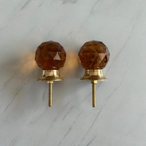 Op de afbeelding: Twee amberkleurige, gefacetteerde glazen knoppen met een bolvorm. Elke knop is voorzien van een gepolijste messingkleurige metalen voet en een metalen draadeind aan de onderkant voor installatie, wat zorgt voor een stevig bevestigingspunt.