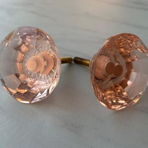 May include: Two round, faceted cabinet knobs made of translucent, pale pink glass. Each knob features a multi-faceted surface that catches the light and is attached to a short, threaded brass-colored metal screw extending from the back center.