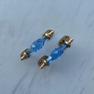 May include: Two drawer pulls featuring faceted, translucent blue glass centres shaped like elongated diamonds. Each pull is secured by metallic, brass-toned hardware at both ends, including mounting plates with visible screw holes for attachment.