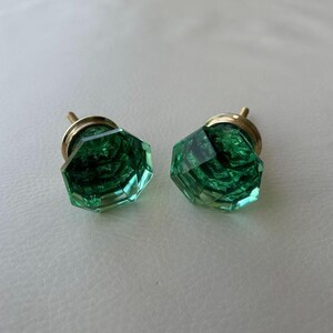 May include: Two emerald green glass cabinet knobs with a faceted, octagonal shape. Each knob features a polished brass-toned metal base and a translucent, multi-faceted surface that reflects light, creating a clear and geometric appearance.