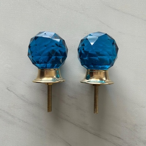 Puede incluir: Dos pomos para armarios con cabezas esféricas facetadas de vidrio azul translúcido, fijadas sobre bases de metal dorado pulido. Cada pomo incluye un tornillo metálico roscado que sobresale de la base para facilitar su instalación.