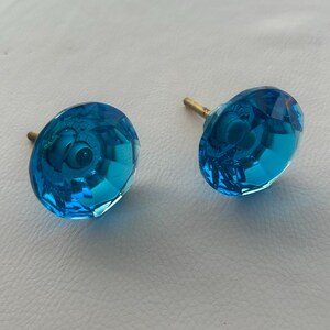 May include: Two faceted, turquoise blue crystal cabinet knobs with brass screw posts. The knobs have a rounded, geometric design, reflecting light. These decorative knobs are suitable for furniture or cabinet upgrades.