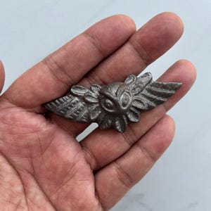 May include: A dark gray metal owl pin with outstretched wings. The owl has detailed feathers and floral accents. The pin is held in a hand against a white background.