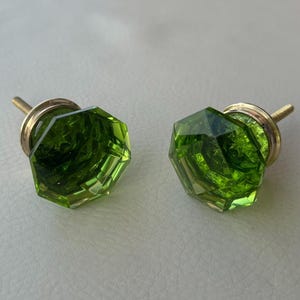 Może przedstawiać: Dwa fasetowane, zielone szklane gałki do szafek z mosiężnym osprzętem. Gałki mają geometryczny kształt i żywy, przezroczysty kolor. Nadają się do projektów mebli i dekoracji wnętrz.