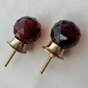 Könnte beinhalten: Zwei facettierte, dunkelbernsteinfarbene Kristall-Schrankknöpfe mit messingfarbenen Sockeln und Gewindestiften. Die Knöpfe haben ein geometrisches, vielseitiges Design. Geeignet für die Möbelrestaurierung.