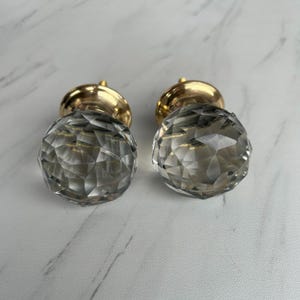 Può includere: Due pomelli rotondi per mobili con cime in vetro trasparente sfaccettato che creano un aspetto geometrico simile a un diamante. Ogni pomello è fissato a una base in metallo color oro lucido, offrendo un contrasto riflettente e metallico.