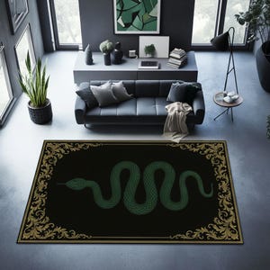 Pode incluir: Um tapete retangular preto com um desenho de cobra verde no centro. A cobra é enrolada e detalhada. O tapete tem uma borda dourada ornamentada e uma moldura dourada. O tapete está colocado sobre um chão cinzento numa sala com um sofá e plantas.