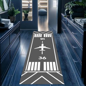 May include: A grey and white airplane runway runner rug with the numbers 18 and 36 printed on it. The rug has a white airplane silhouette in the centre.