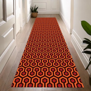 May include: A long, rectangular runner rug with a repeating geometric pattern of orange, red, and black hexagons. The rug is placed in a hallway with light-colored wooden floors and white walls. The rug's design is a bold statement.