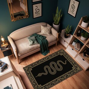 Op de afbeelding: Een zwart rechthoekig vloerkleed met een gouden slangenontwerp en een sierlijke rand. Het vloerkleed ligt in een kamer met een beige bank, een groene plaid en houten planken. De muren zijn donkerblauwgroen geschilderd.