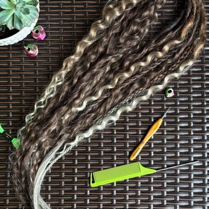 May include: A bundle of brown, blonde, and gray dreadlocks with a wavy texture. A green comb, a crochet hook, and decorative beads are next to the dreadlocks. A succulent plant in a white pot is in the background.
