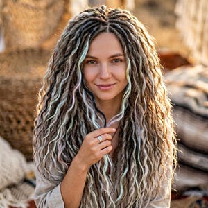 May include: Long, wavy synthetic dreadlocks in shades of brown, gray, and white, featuring thin strands of pastel mint green and soft pink throughout. The hair extensions are styled in a voluminous, textured manner with a center part.
