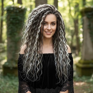 May include: A woman with long, wavy hair in shades of gray and brown, styled in dreadlocks. She wears a black off-the-shoulder top with lace detailing. The background is a blurred forest scene with green foliage.