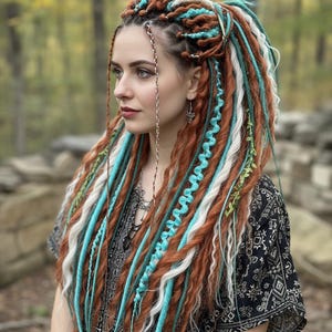 Puede incluir: Una mujer con largas y coloridas rastas. Las rastas son una mezcla de marrón, blanco y turquesa, con algunos acentos verdes. Lleva una blusa negra estampada y joyas de plata. El fondo es un bosque borroso.