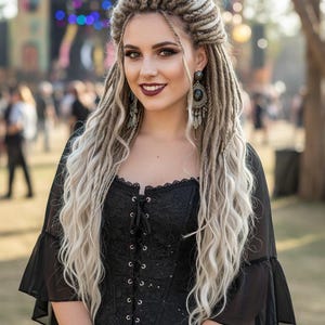 May include: A woman with long, blonde dreadlocks and dark lipstick smiles. She wears a black lace corset top with a lace-up front and bell sleeves. Silver bracelets and earrings complete the look. The background is blurred, suggesting an outdoor event.
