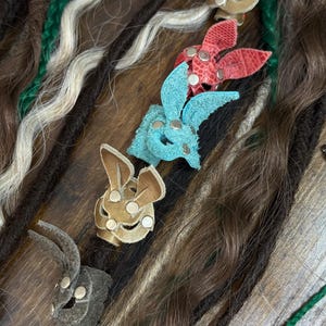 May include: An assortment of dreadlock accessories, including leather bunny-shaped hair clips in various colors like tan, red, and blue. The dreadlocks are a mix of brown, black, green, and white.