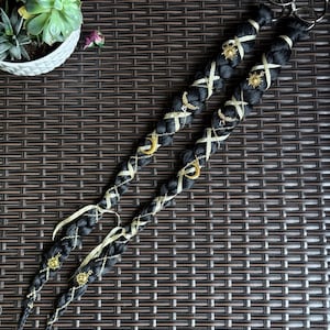 May include: Two black braided hair extensions with gold accents. The braids are adorned with gold ribbons, charms, and sun and moon pendants. Silver rings are attached at the top. The extensions are displayed on a brown woven surface.