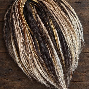May include: A set of synthetic hair extensions featuring a mix of dark brown, light brown, and blonde strands. The hair is styled into a combination of textured dreadlocks and thin, smooth braids, all secured together at the top with a green tie.
