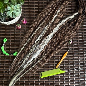 Puede incluir: Dreadlocks marrones y blancas con textura ondulada. Las rastas están dispuestas sobre una superficie tejida marrón, con un peine verde, un ganchillo y otros accesorios cerca. Una pequeña planta suculenta está en el fondo.