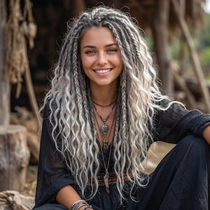 Puede incluir: Una persona con cabello largo, ondulado y de color gris plateado, peinado con múltiples trenzas. Viste una prenda negra de manga larga, collares superpuestos con colgantes oscuros y pulseras apiladas en la muñeca, sonriendo.