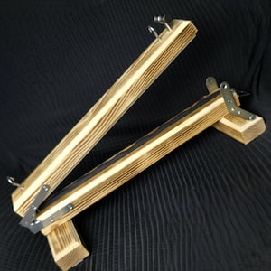 May include: A wooden embroidery hoop stand with a natural wood grain finish. The stand features a hinged design with metal hardware and is designed to hold an embroidery hoop. The stand is angled and set against a dark background.