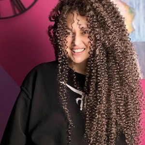 May include: A person with long, curly brown hair smiles, wearing a black sweatshirt with a white design. The background is a pink wall with a black clock and a painting.