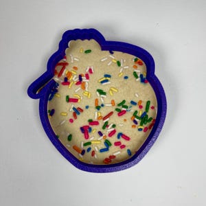 May include: Purple plastic cookie cutter shaped like a sheep with a cookie inside. The cookie is covered in colorful sprinkles.