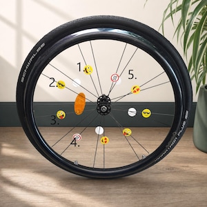 Può includere: Una ruota di bicicletta nera con pneumatico Schwalbe Marathon Plus, caratterizzata da diversi adesivi circolari applicati ai raggi. Gli adesivi includono varie faccine emoji gialle, cerchi bianchi e segnali rossi di limite di velocità.